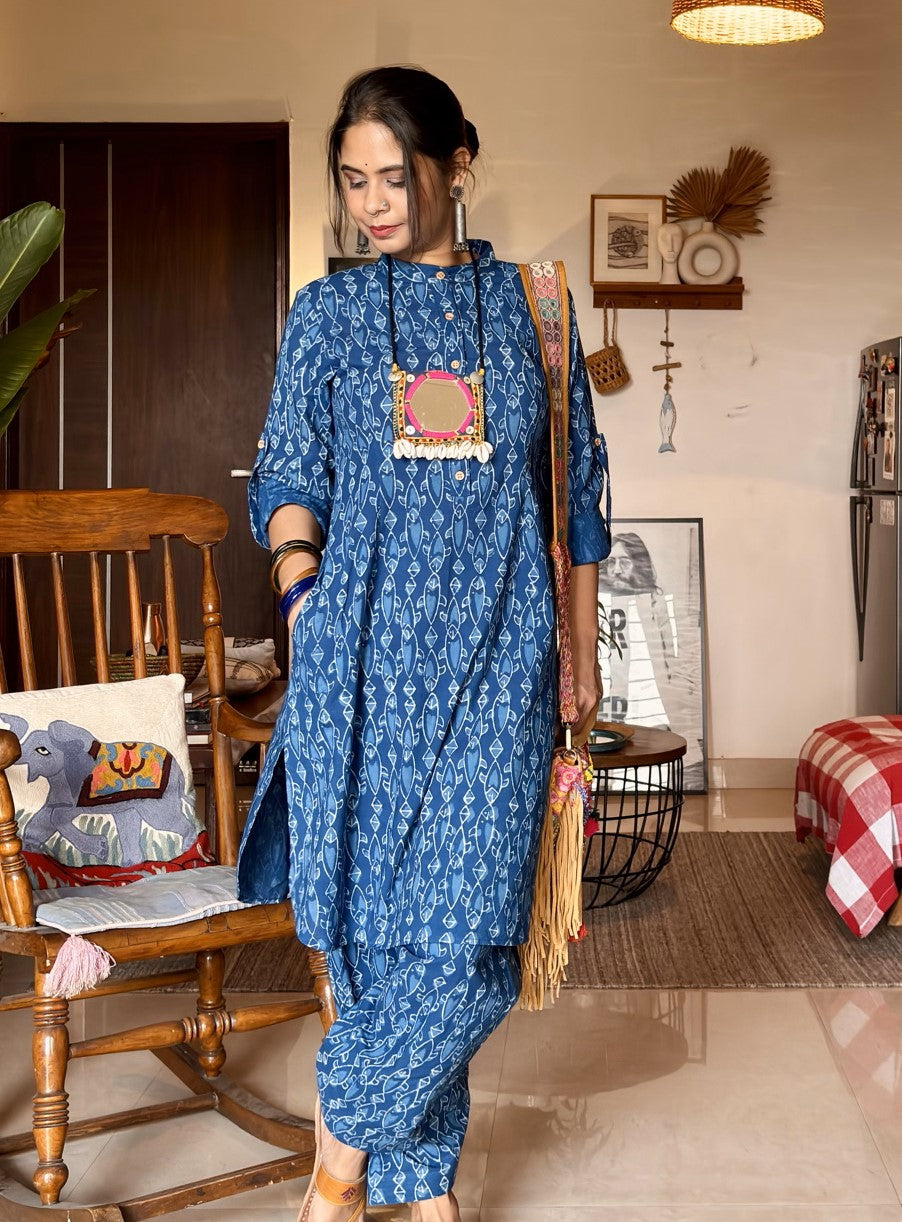 A person wearing a blue kurta and pant coord set with a fish print design, standing in a room with wooden furniture and decorative items on the walls.