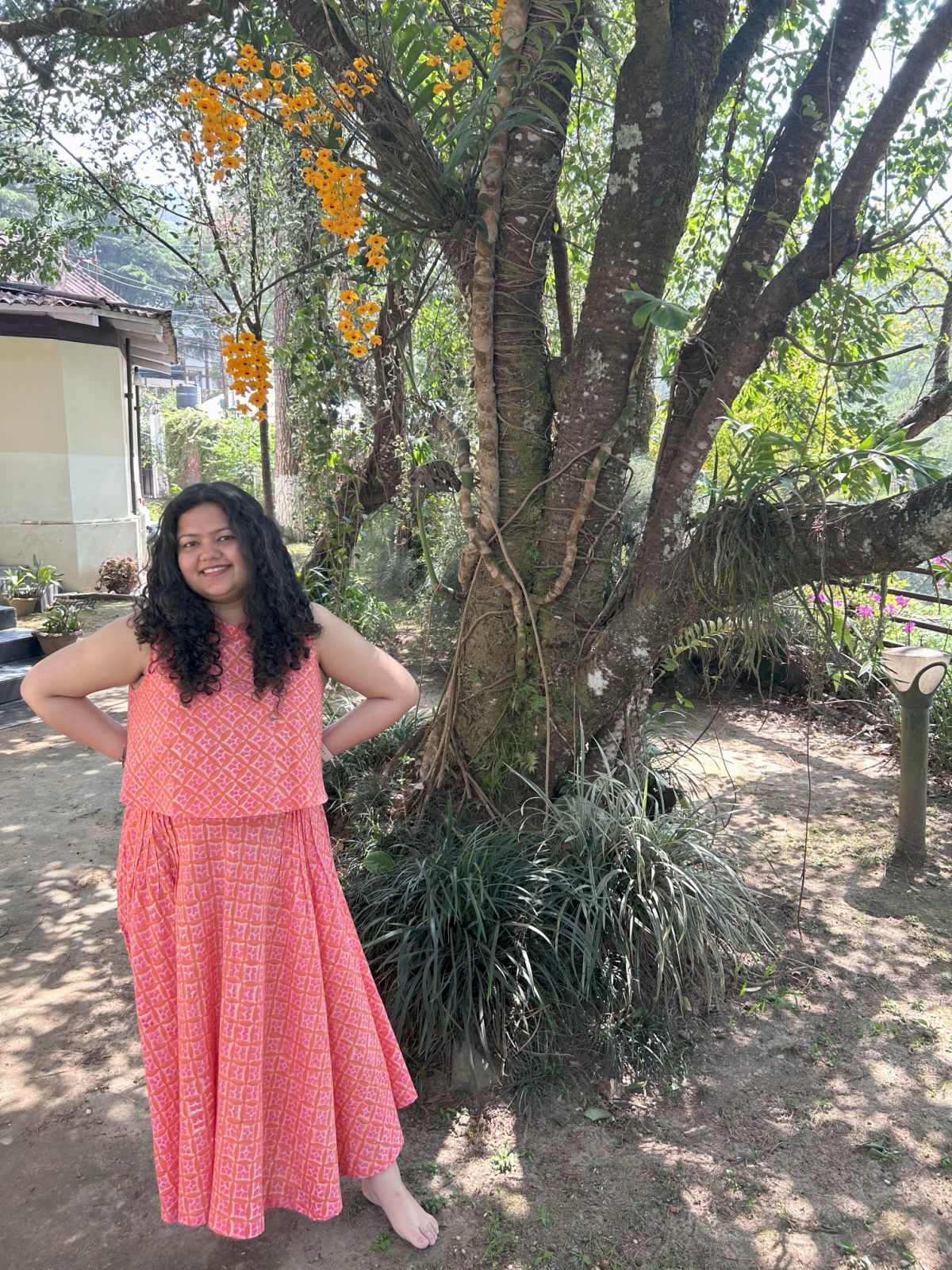Woman in a pink dress standing next to a tree with orange flowers in a garden setting.