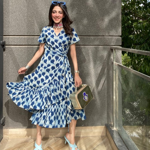 A person wearing a blue floral frill dress with short sleeves and a tie up belt, standing near a glass railing.