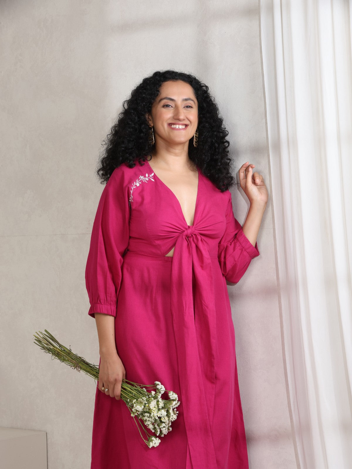 A woman standing by a window, wearing a pink maxi dress with a long front knot and floral embroidery on the shoulder, holding a bouquet of dried flowers.