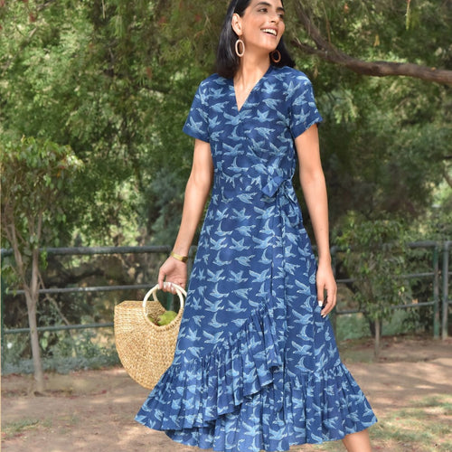 A woman standing outdoors in a blue indigo dress with a leaf pattern. She is wearing a tie-up belt and the dress features short sleeves and frills at the hem.